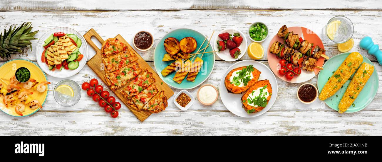 Summer BBQ grill table scene over a white wood banner background. Grilled flatbread, chicken and shrimp skewers, stuffed sweet potato, corn, fruit and Stock Photo