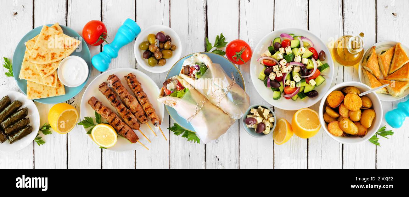 Greek food table scene, top view on a white wood banner background. Variety of items including gyros wraps, souvlaki, salad, spanakopita, dolmades, pi Stock Photo