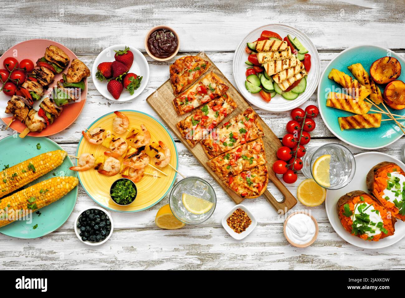 Summer BBQ grill table scene over a white wood background. Chicken and shrimp skewers, flatbread, stuffed sweet potato, grilled fruit, corn and salad. Stock Photo