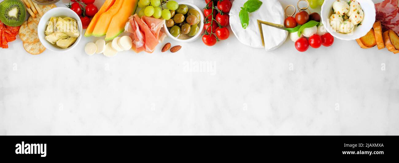 Italian theme charcuterie top border against a white marble banner background. Assortment of cheese, meat and fruit appetizers. Above view with copy s Stock Photo