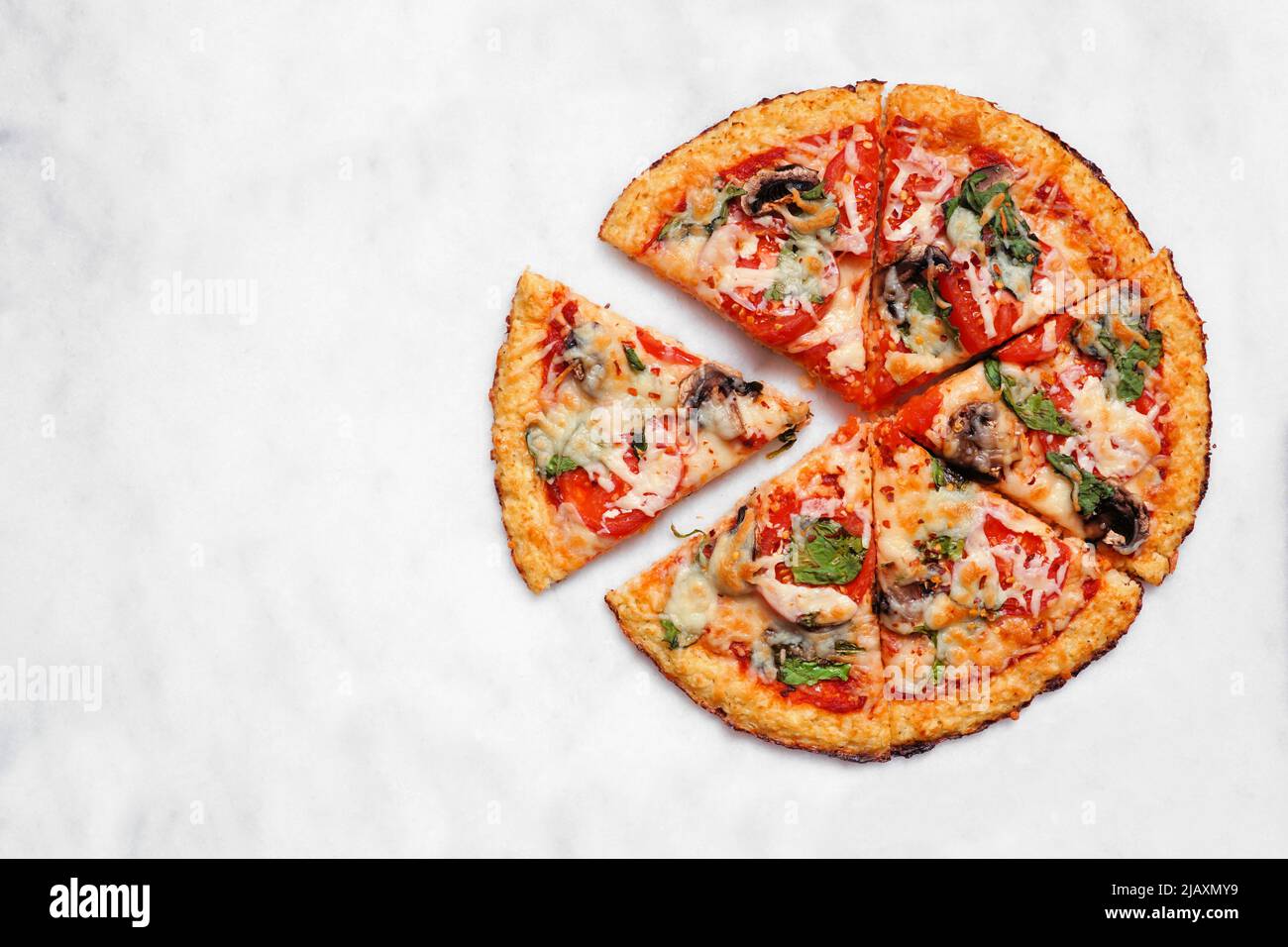 Healthy, gluten free cauliflower crust pizza with tomatoes, mushrooms and spinach. Overhead view with cut slices on a white marble background. Stock Photo