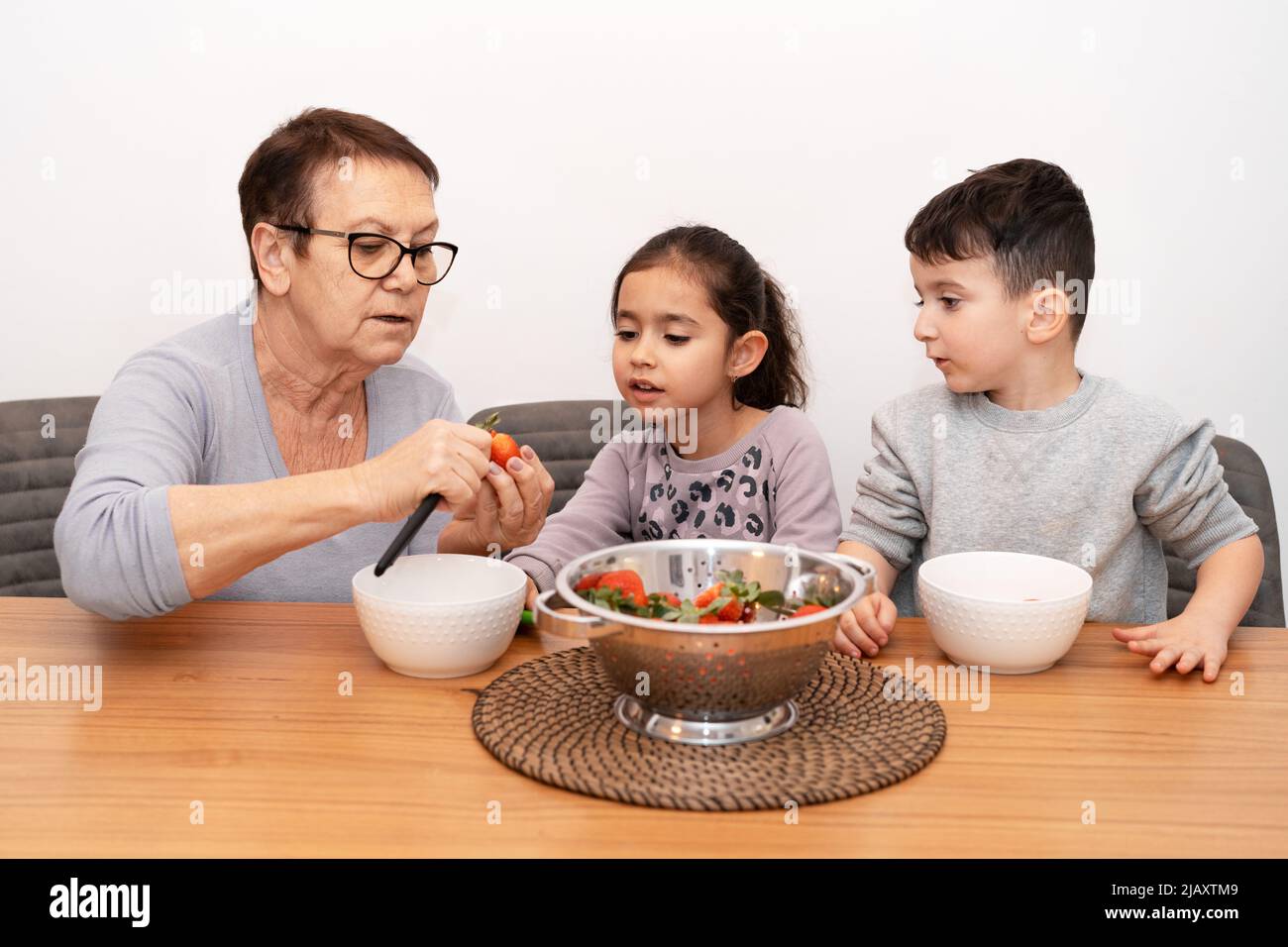 Senior woman and two little children in the kitchen cutting fruit salad. Grandmother and grandchildren cook strawberry jam together. Stock Photo