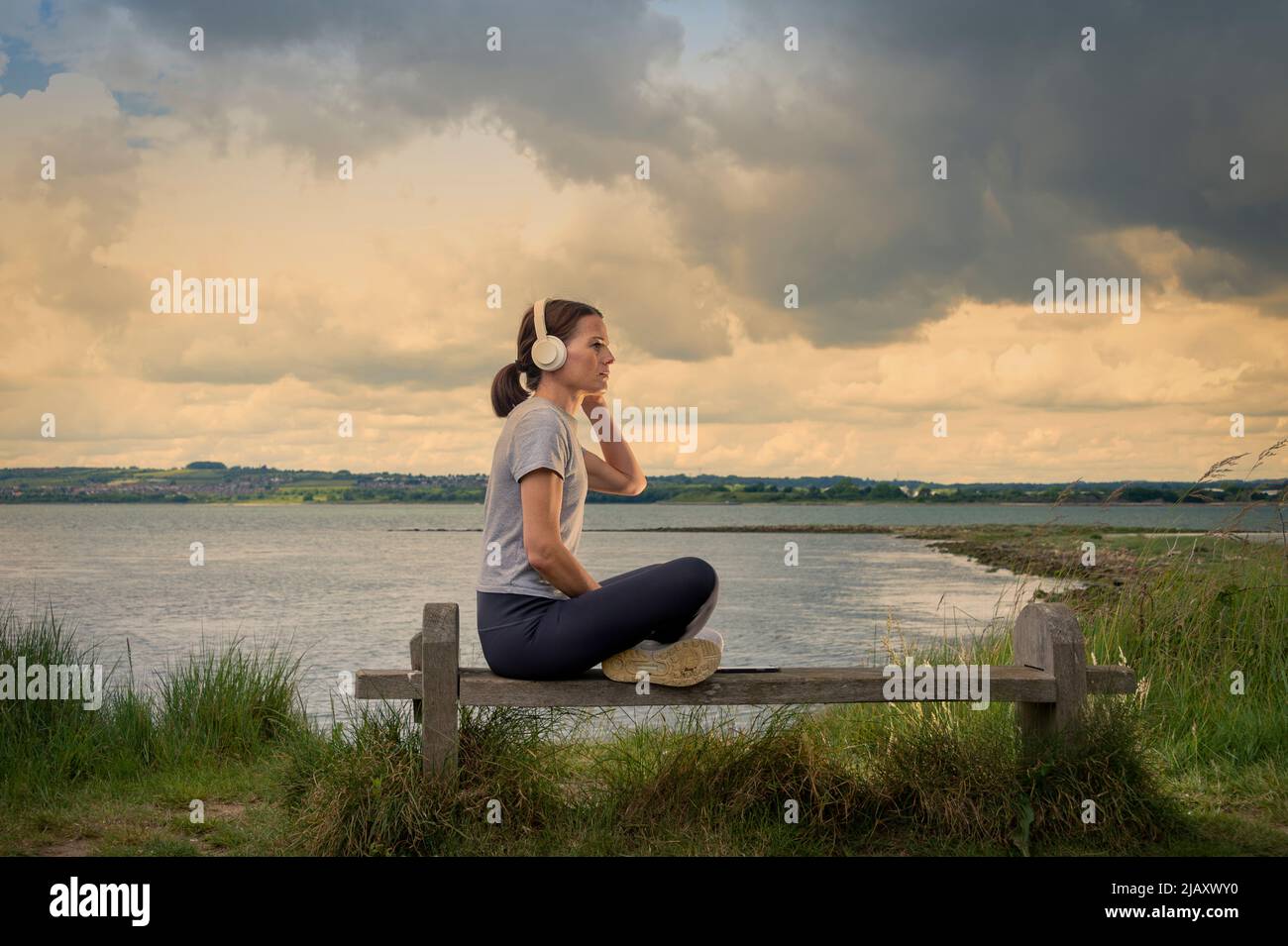 Single woman sitting on a bench beside water listening to music on her headphones. Stock Photo