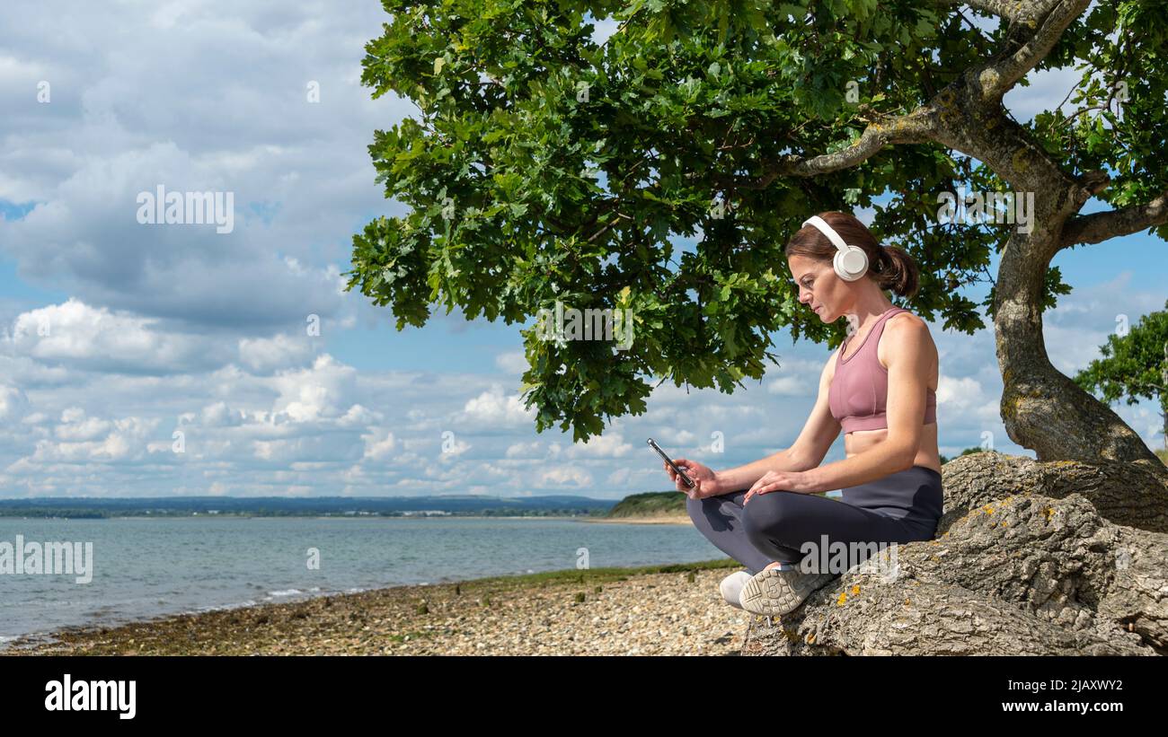 woman sitting under a tree by water listening to podcast or music in the sun. Stock Photo