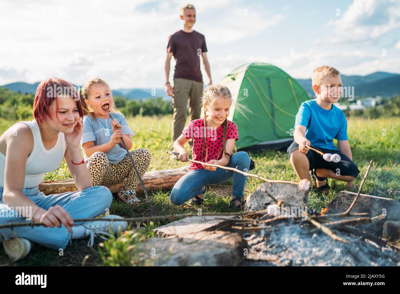 Boys and girls friends kids cheerfully laughing and roasting marshmallows on sticks over campfire flame near the green tent. Outdoor active time spend Stock Photo