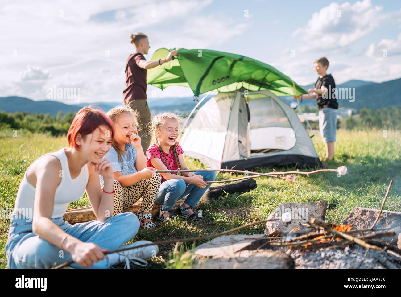 Three sisters cheerfully laughing and roasting marshmallows and candies on sticks over campfire flame while two brothers set up the green tent. Happy Stock Photo