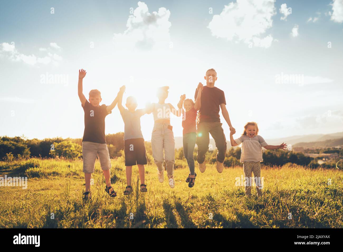 Six kids brothers and sisters teenagers and little kids funny jumping holding hands in hands on the green grass meadow with an evening sunset backgrou Stock Photo