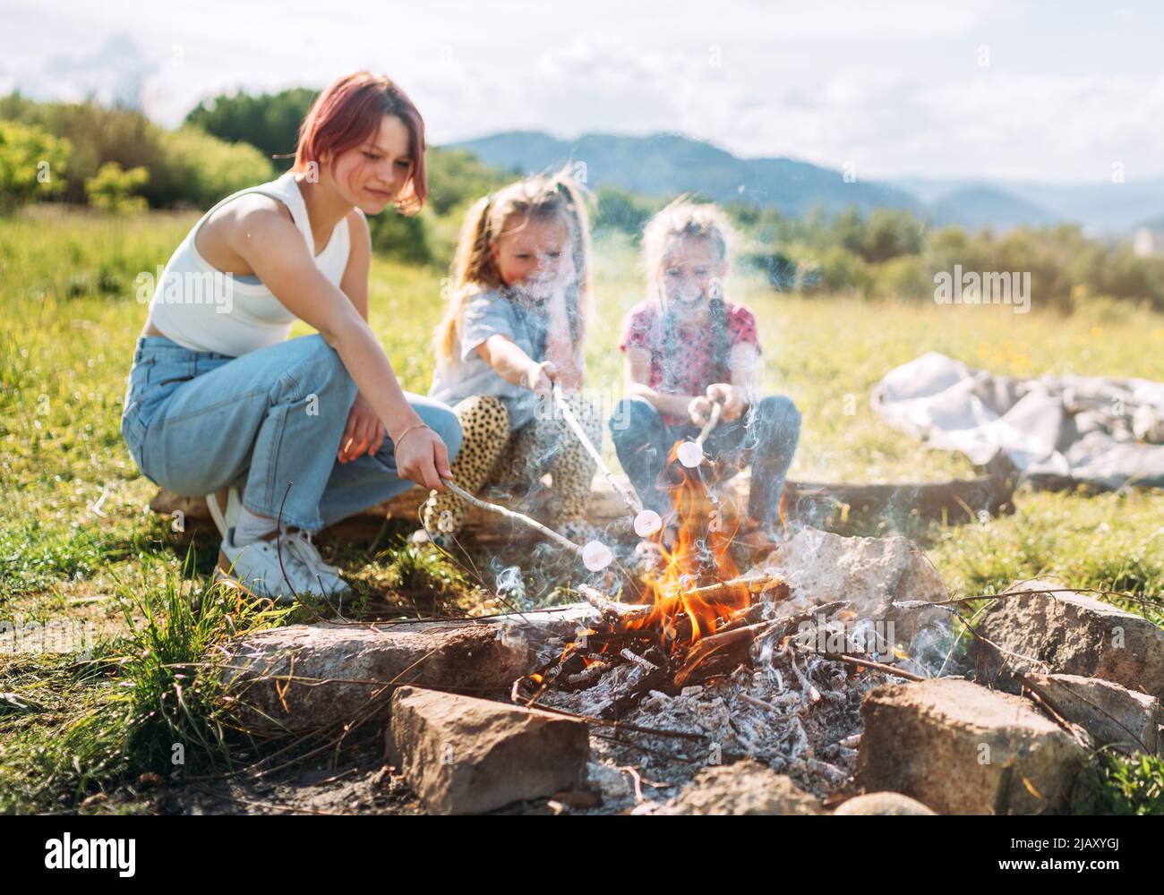 Three sisters cheerfully smiling while they roasting a  marshmallows candies on the sticks over the campfire flame. Happy family or outdoor picnic act Stock Photo