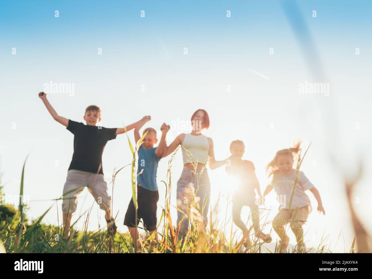 Selective focus brothers and sisters teenagers and little kids jumping holding hands in hands on the green grass meadow with an evening sunset backgro Stock Photo