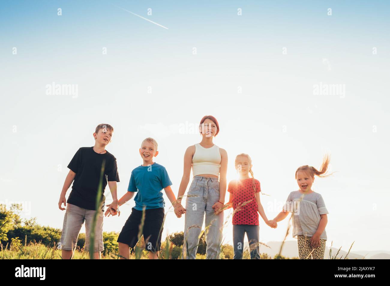 Five kids brothers and sisters teenagers and little kids walking on the green grass meadow with evening sunset background light holding hands in hands Stock Photo