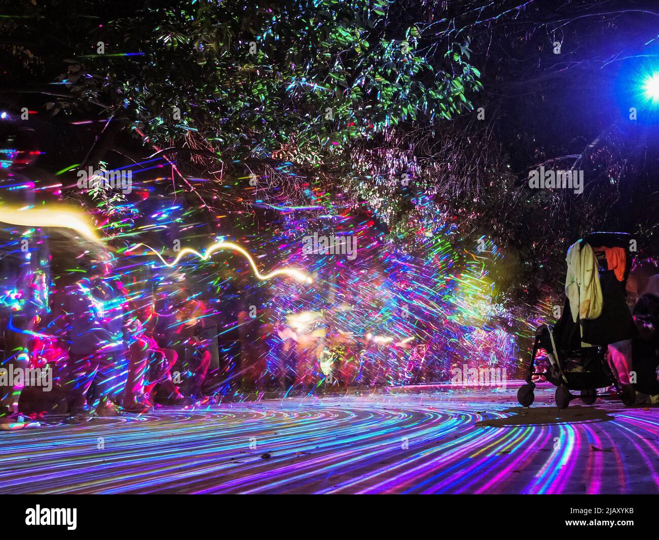 Bright multi-colored lines glow on the alley in the night park creating pattern on the defocused background of people walking. Play of light and shado Stock Photo