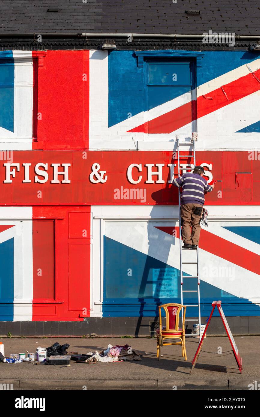 Cradley Heath, West Midlands, UK. 1st June, 2022. Chip shop proprietor Warren Rudge puts the finishing touches onto the Union Jackflag he and his son James have painted on their fish and chip shop in Cradley Heath, West Midlands. The painted flag covers the whole of Ivan's Fish and Chips frontage including the windows, and has taken all day to complete. 'Ivan's Fish and Chips has been serving fish and chips for 65 years - not quite as long as Her Majesty though. But I have heard that The Queen is partial to fish and chips.' says Warren. Credit: Peter Lopeman/Alamy Live News Stock Photo