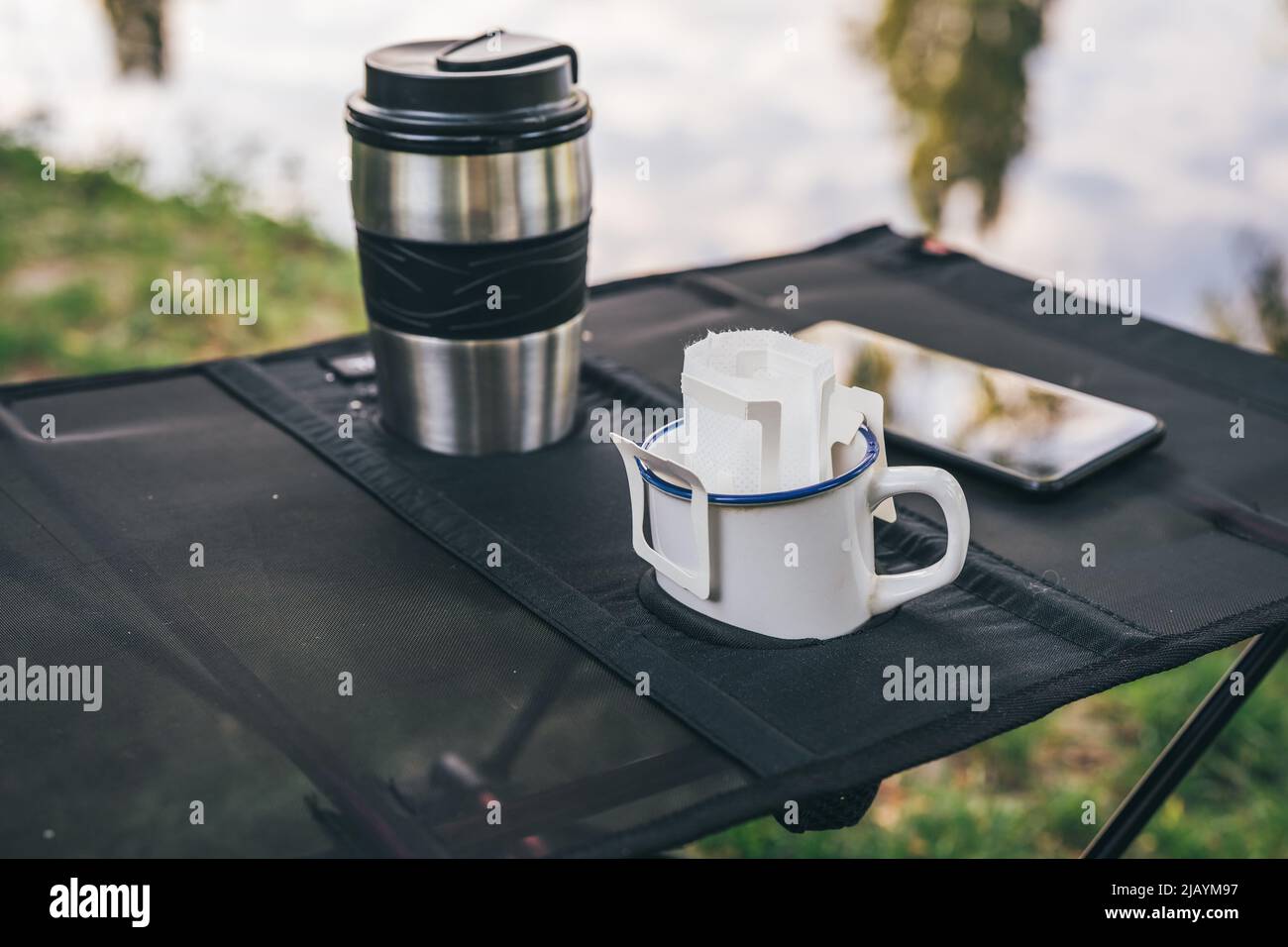 Trendy convenient paper drip coffee bag in metal cup on camping table outdoors. Making freshly brewed coffee in nature Stock Photo