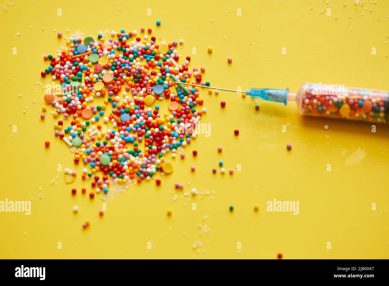 Close-up of splattering sweet sprinkles and needle of medical syringe on bright yellow background Stock Photo