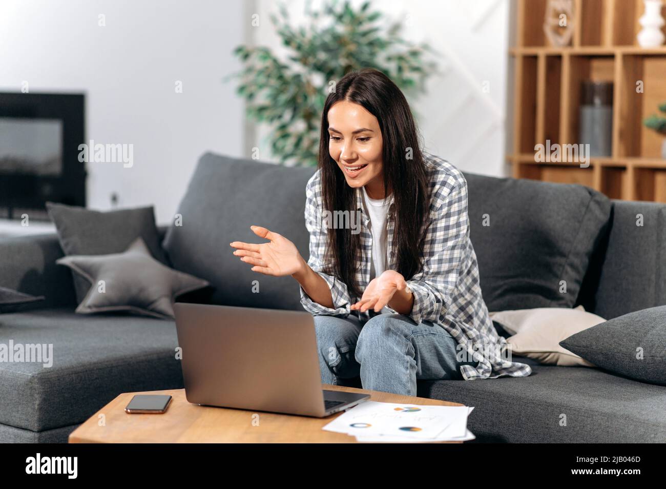 Friendly caucasian successful brunette woman, freelancer or small business owner, sit on a sofa in the living room, using a laptop to work from home, communicate with colleague by video call, smiles Stock Photo