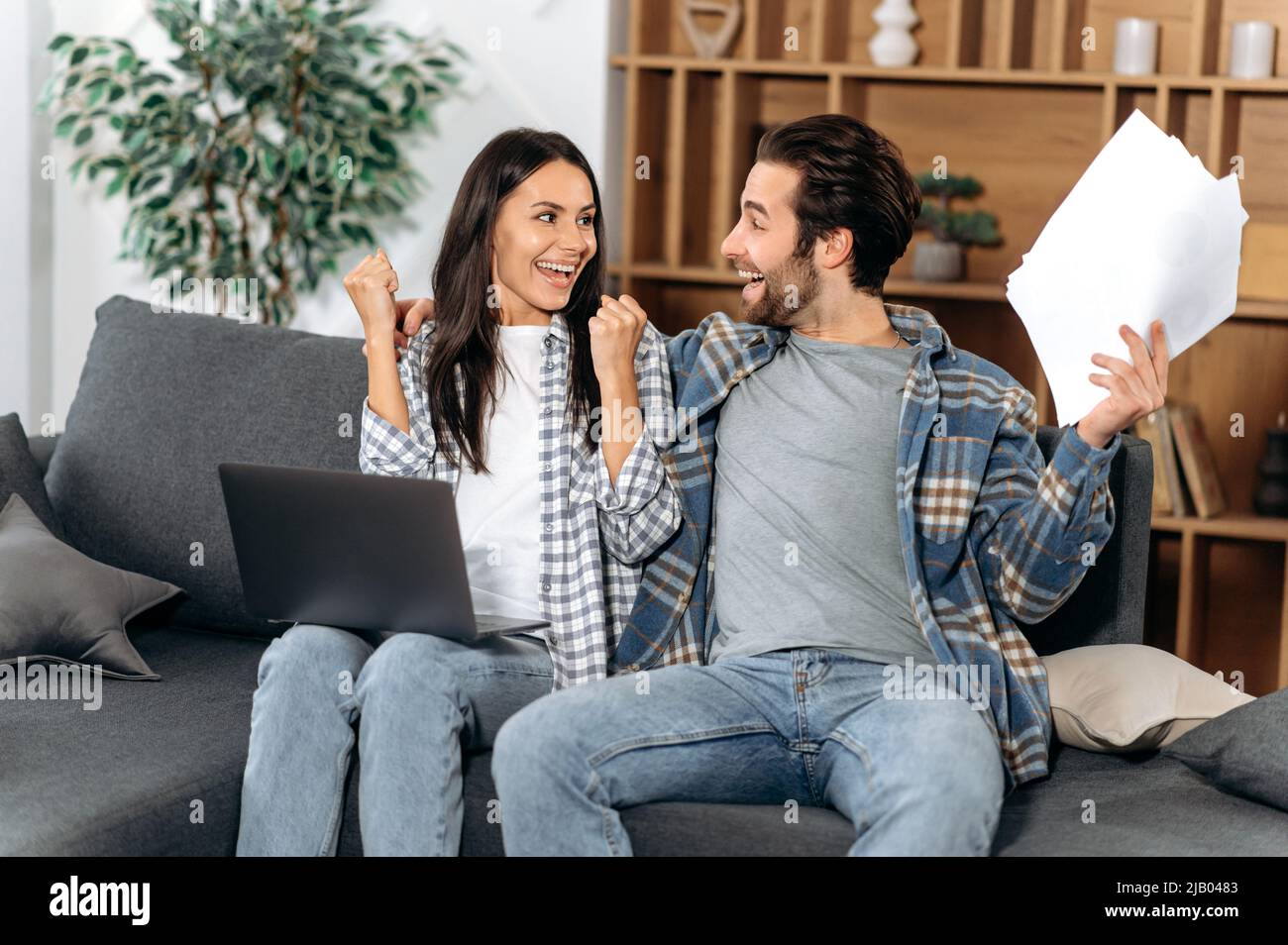 Happy family couple, in stylish clothes, sit on a sofa in the living room, use a laptop and documents, rejoice in big profits or buying real estate, gesticulate with hands, look at each other, smiling Stock Photo