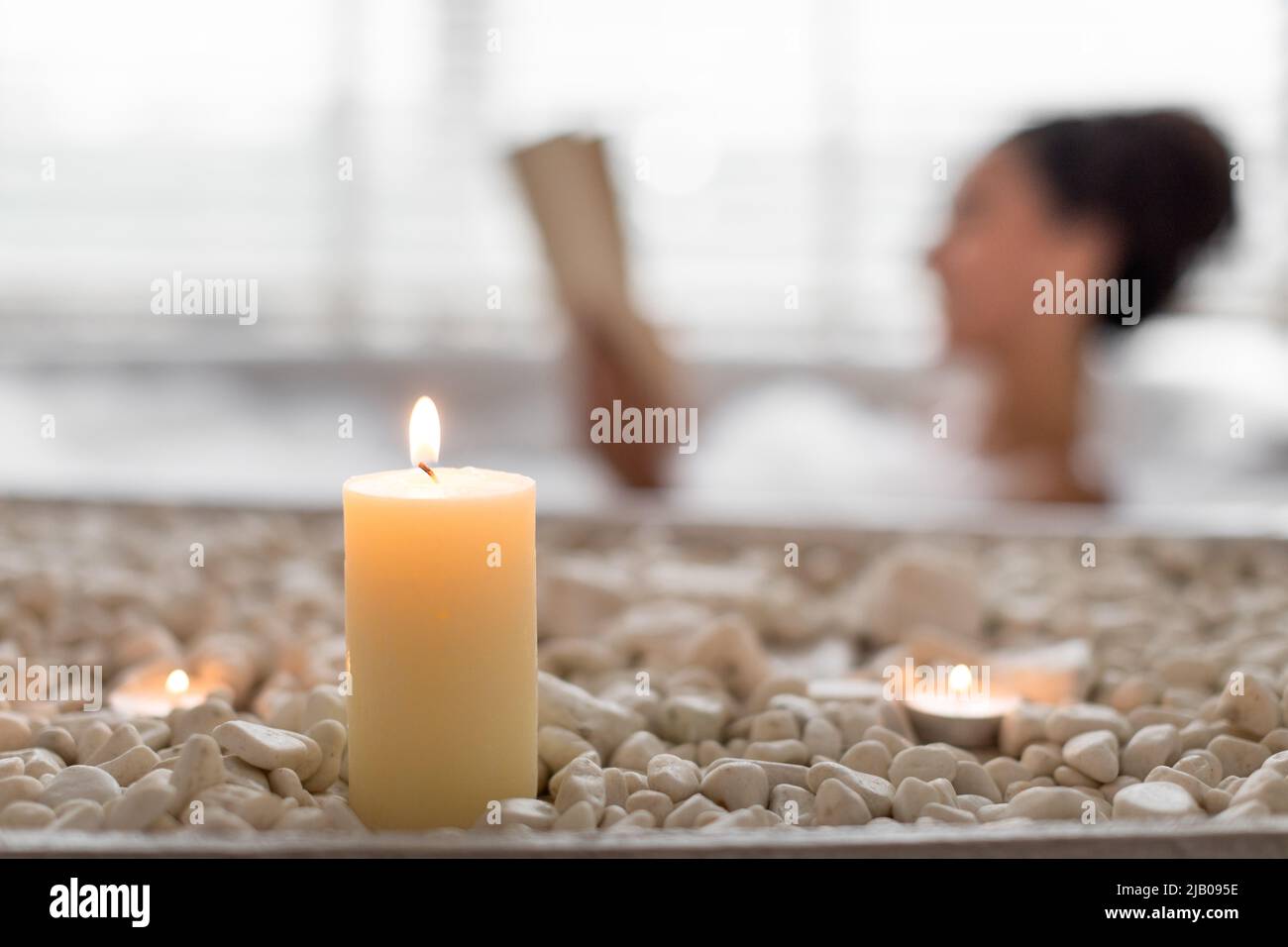 Young woman lying in foamy bath, reading book in relaxing atmosphere, selective focus on candles and stones Stock Photo