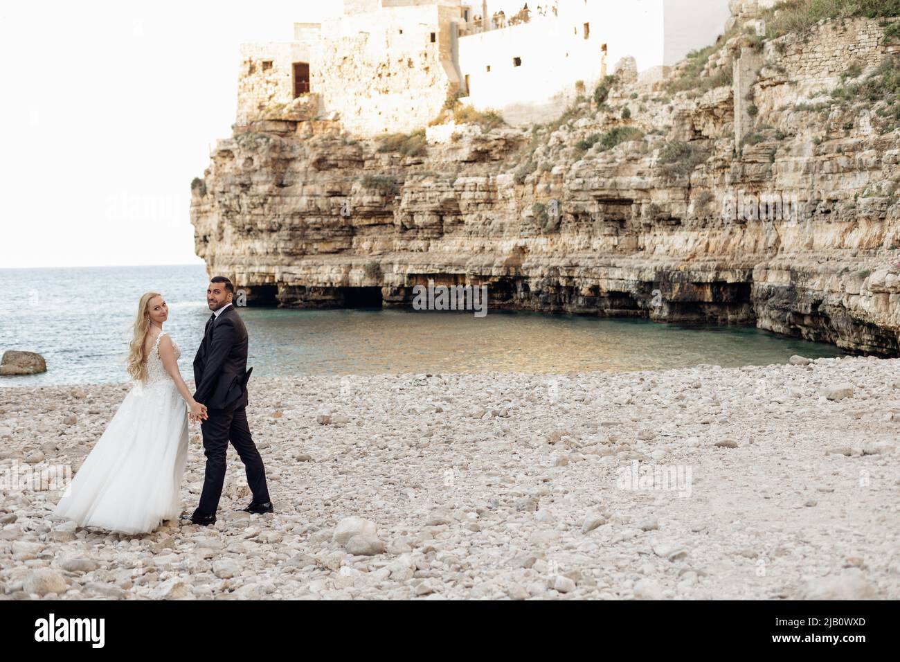 Married couple in wedding dress, official suit standing on the beach near sea. Travelling abroad in Italy, new beginning Stock Photo
