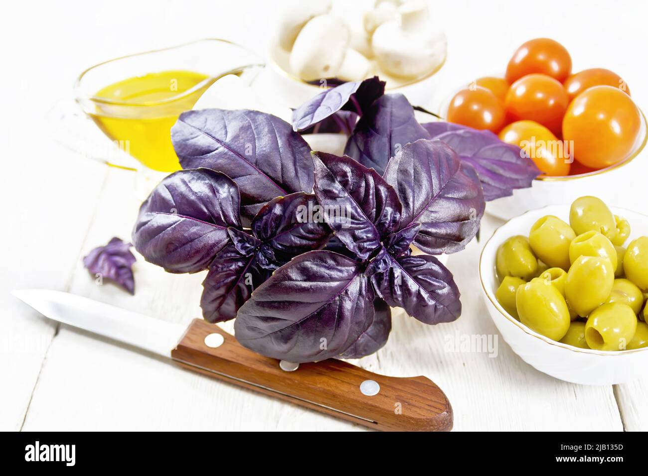 Fresh purple basil in a mortar, olives, tomatoes and champignons in bowls, vegetable oil in gravy boat and a knife on white wooden board background Stock Photo