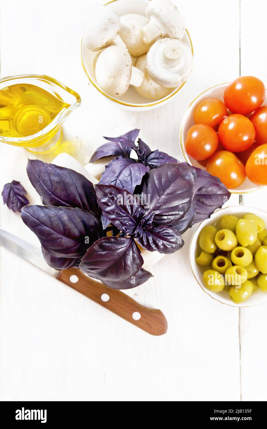 Fresh purple basil in a mortar, olives, tomatoes and champignons in bowls, vegetable oil in gravy boat and a knife on wooden board background from abo Stock Photo