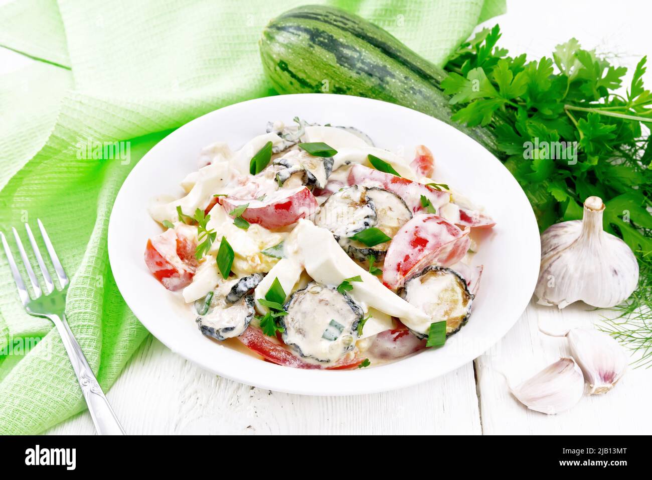 Salad with fried zucchini, boiled egg, fresh tomato and garlic, dressed with mayonnaise in plate, napkin and parsley on white wooden board background Stock Photo