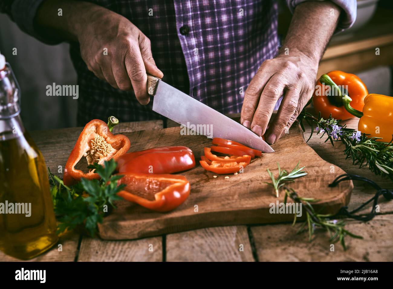 High angle of crop unrecognizable male chef in checkered shirt cutting fresh ripe red bell pepper on wooden chopping board standing at table with oliv Stock Photo