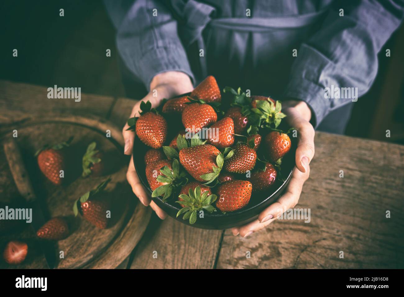 High angle of crop anonymous female in casual dress showing bowl of delicious fresh ripe strawberries standing at wooden table in kitchen in daytime Stock Photo