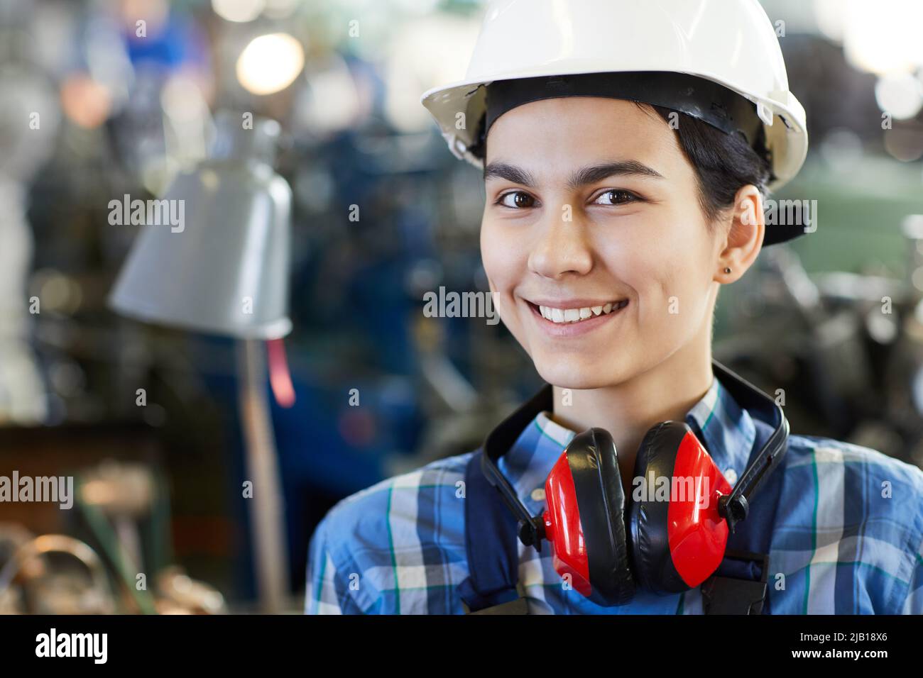 Portrait of smiling beautiful young lady worker in hardhat wearing red ear protectors on neck, woman in industry concept Stock Photo