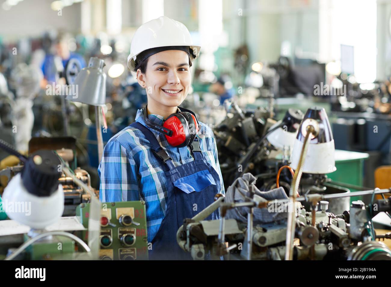 Positive confident attractive masculine lady engineer in hard hat standing at machine for producing clock movements and looking at camera, she working Stock Photo