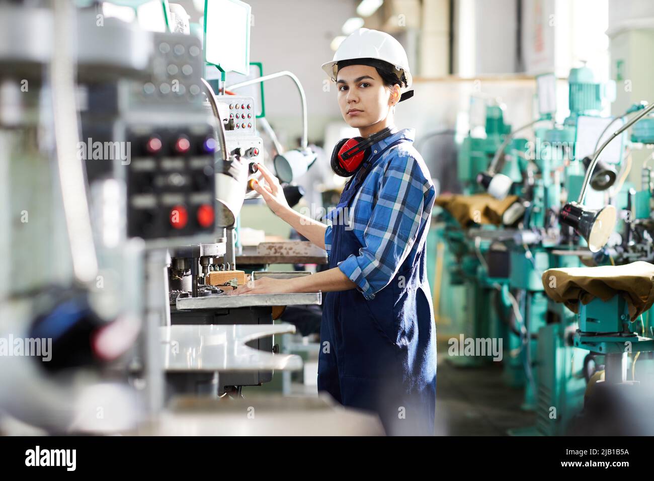 Serious thoughtful woman engineer in uniform standing at industrial machine and producing small gear for wristwatch, she working at production line Stock Photo