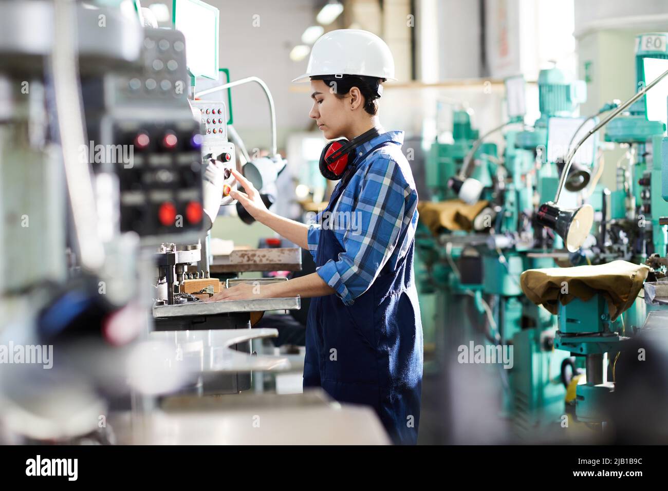 Serious young female engineer in hardhat concentrated on production process pushing button on control panel of industrial machine Stock Photo
