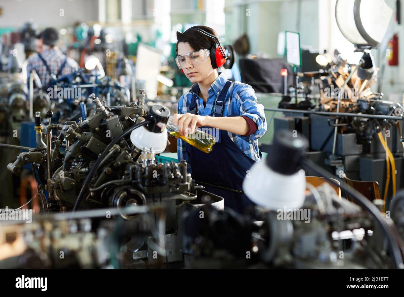 Serious concentrated lady engineer in ear protectors and protective goggles pouring lubricant for machine while working at factory Stock Photo