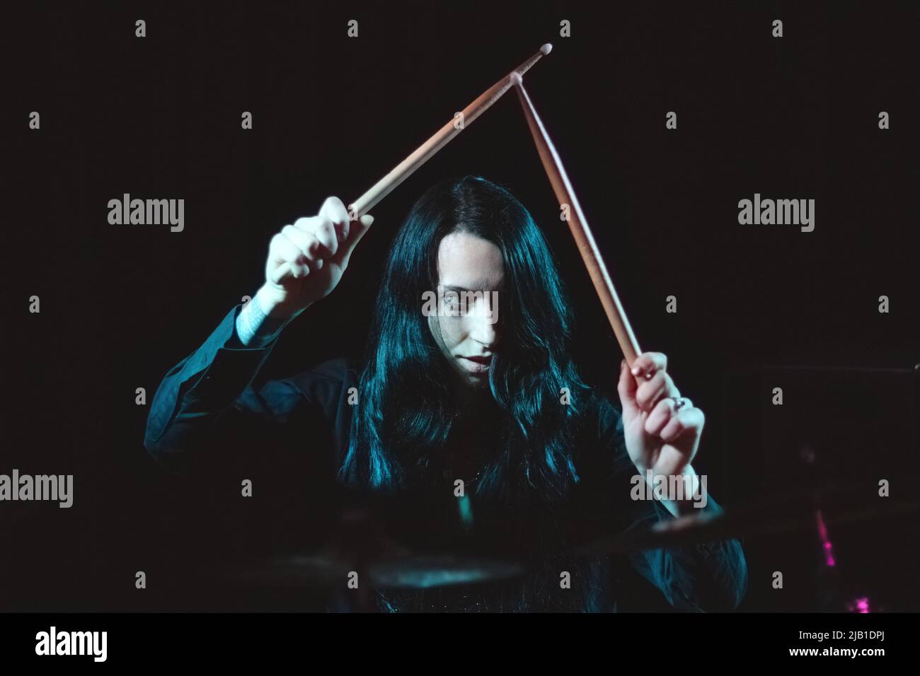 A female metal drummer with long hair holding up drumsticks behind the drumkit against a dark background Stock Photo