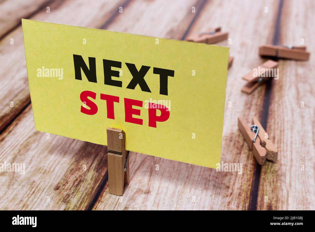 Message card “Next Step” on shabby wooden table. Natural wooden pinch holding yellow paper note. Keep trying to achieve the personal goal. Stock Photo