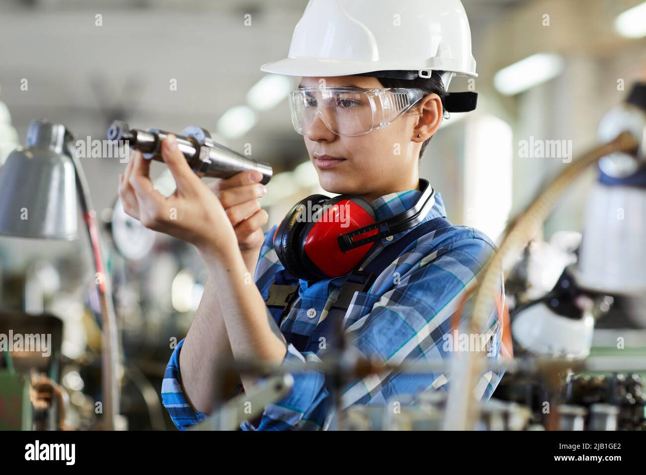 Serious female technical engineer in construction hardhat and safety goggles checking drill bit while repairing machine for producing clock movements Stock Photo
