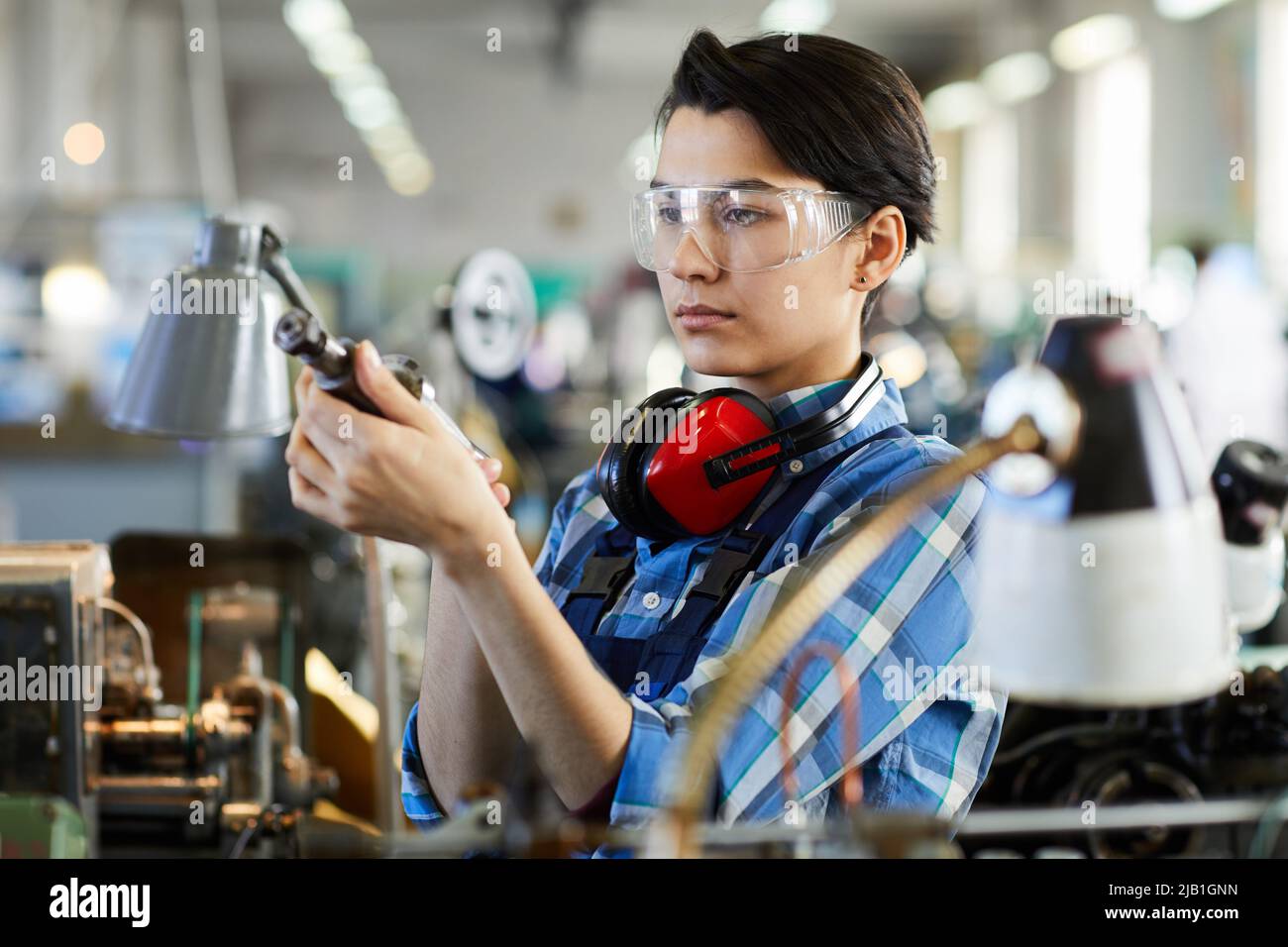 Concentrated young female repairing engineer in safety goggles standing between industrial machines and viewing cutting tool at factory shop Stock Photo