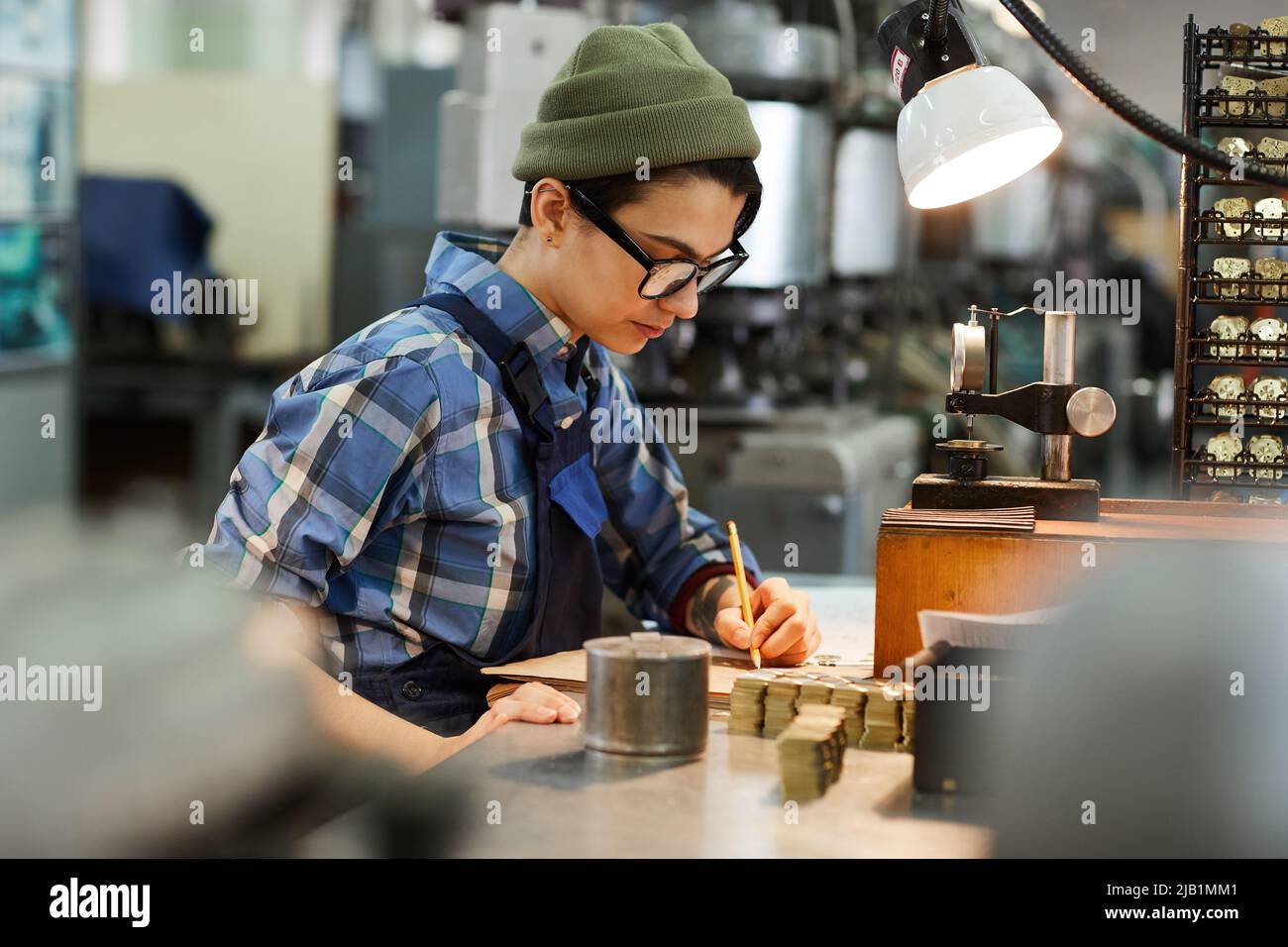 Serious concentrated female watch factory engineer in glasses sitting at desk and viewing notes while analyzing production of gears and cogs Stock Photo