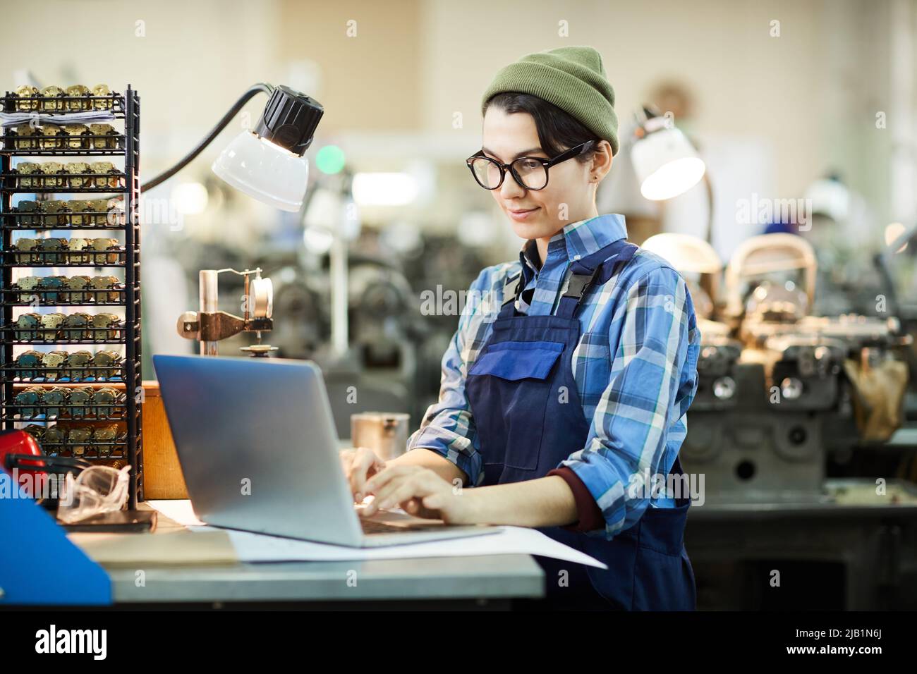 Content busy tomboy engineer in overall standing at table and typing on laptop while composing online guidance at watch factory Stock Photo
