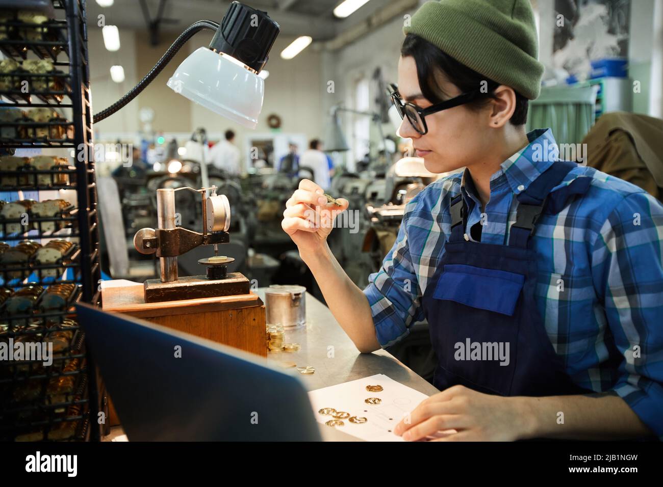 Serious concentrated hipster engineer in beanie hat and glasses sitting at table and checking quality of small details of watches Stock Photo