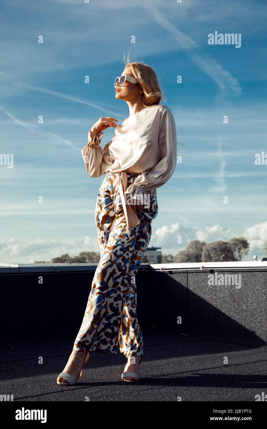 Young woman with long fair hair wearing silk blouse, printed trousers, sunglasses, standing with crossed legs on roof. Stock Photo