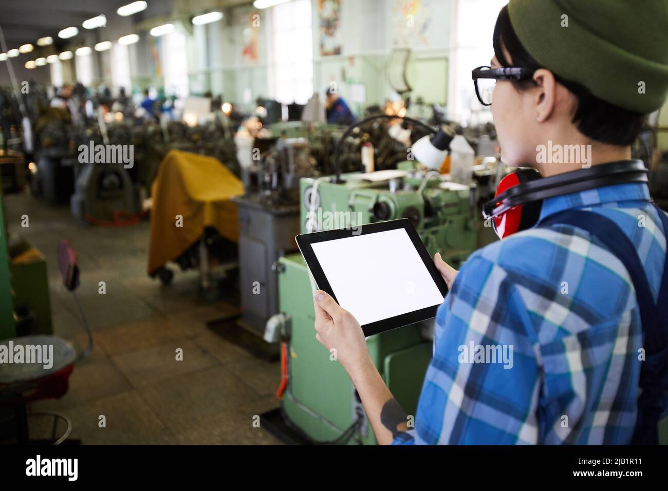 Rear view of pensive woman engineer with ear protectors on neck using digital tablet while inspecting production shop at factory Stock Photo