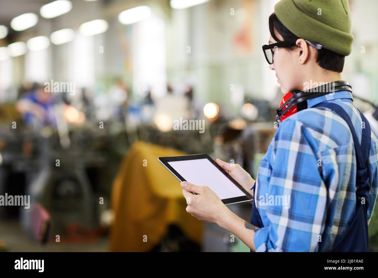 Serious busy lady engineer in hipster hat and glasses standing in industrial shop and using tablet while viewing online notes Stock Photo