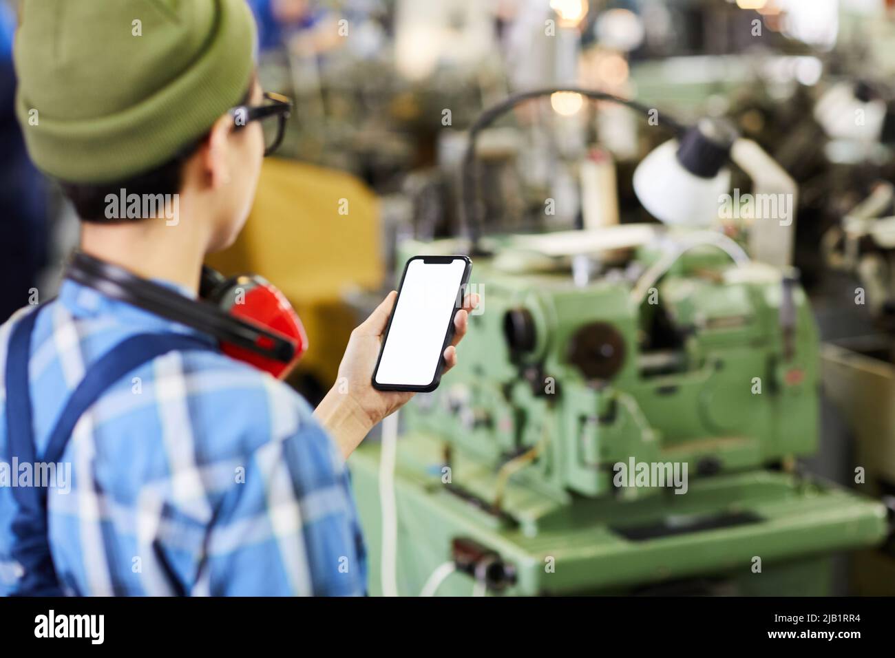 Rear view of lady engineer in beanie hat standing in industrial shop and scanning information using smartphone Stock Photo