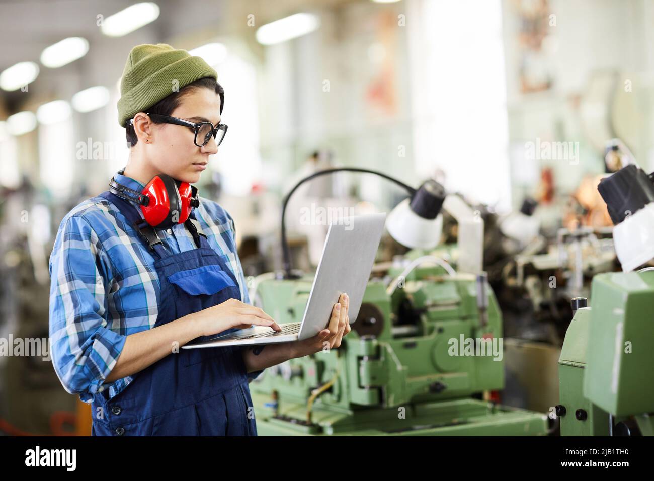 Serious lady engineer in overall standing in factory shop and examining online sketch on laptop Stock Photo