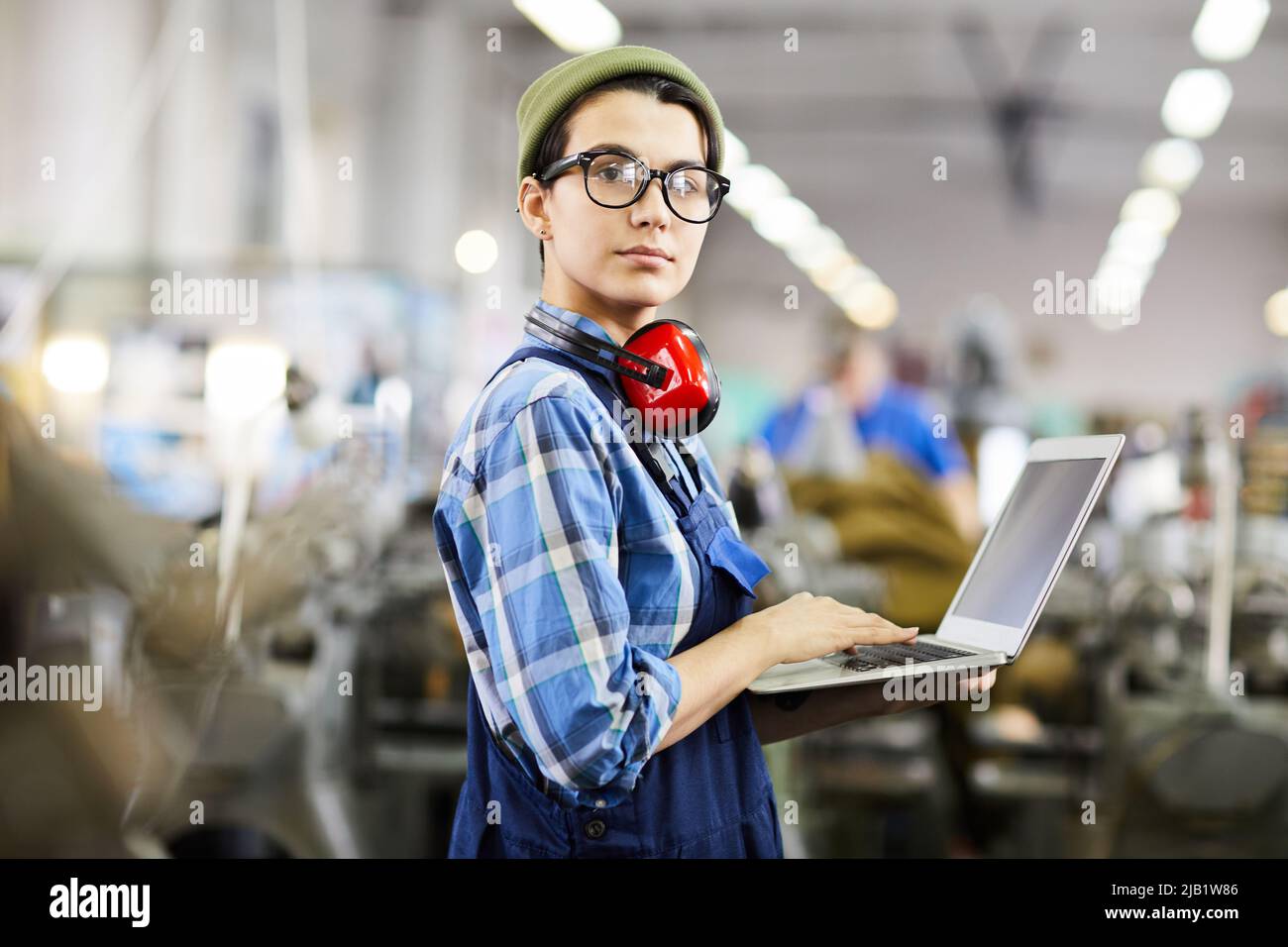 Serious confident female engineer in beanie hat and eyeglasses standing in industrial shop and using laptop while looking at camera, woman in industry Stock Photo