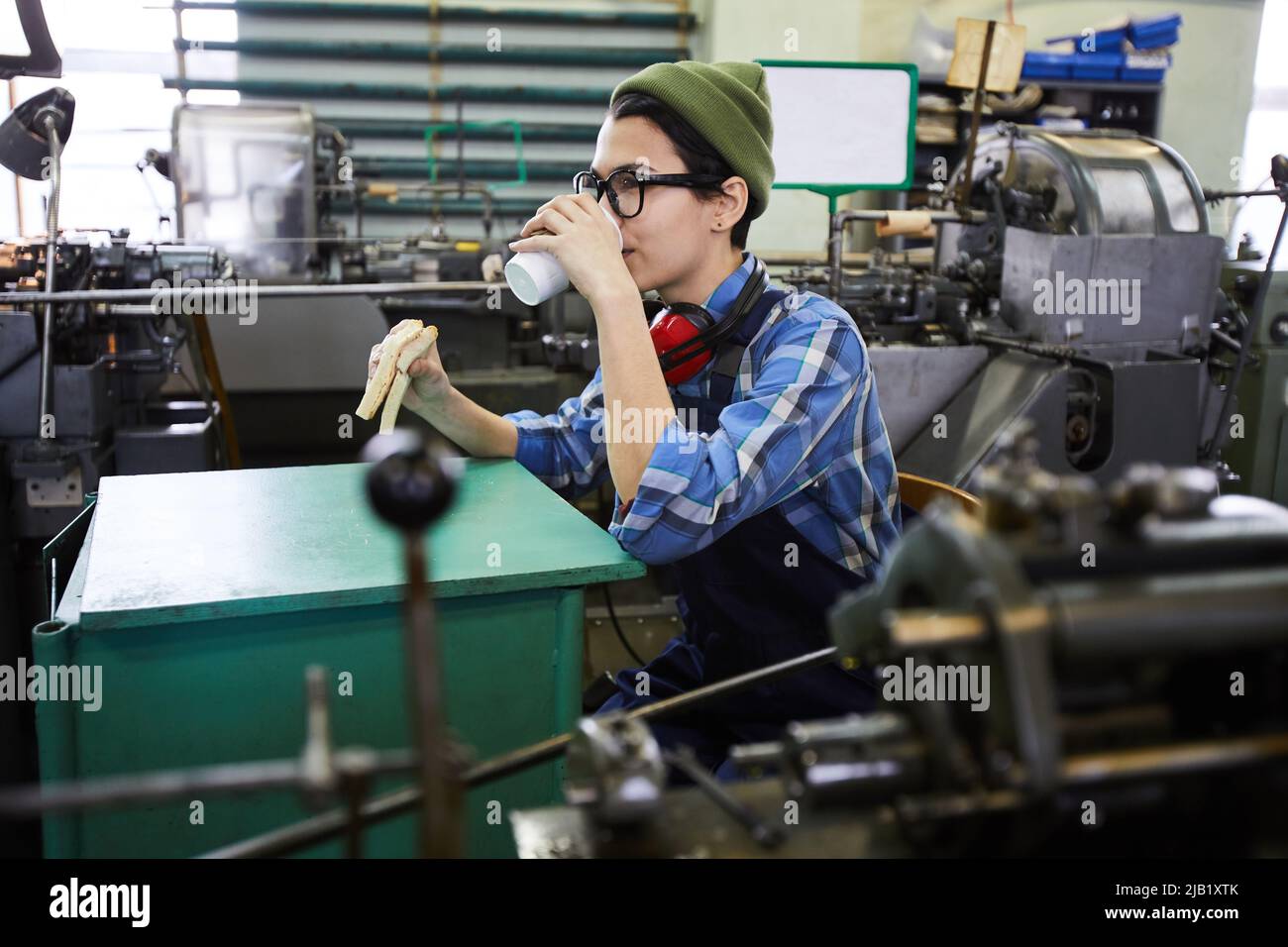Content relaxed young woman in workwear drinking takeout coffee and eating sandwich at factory workplace Stock Photo