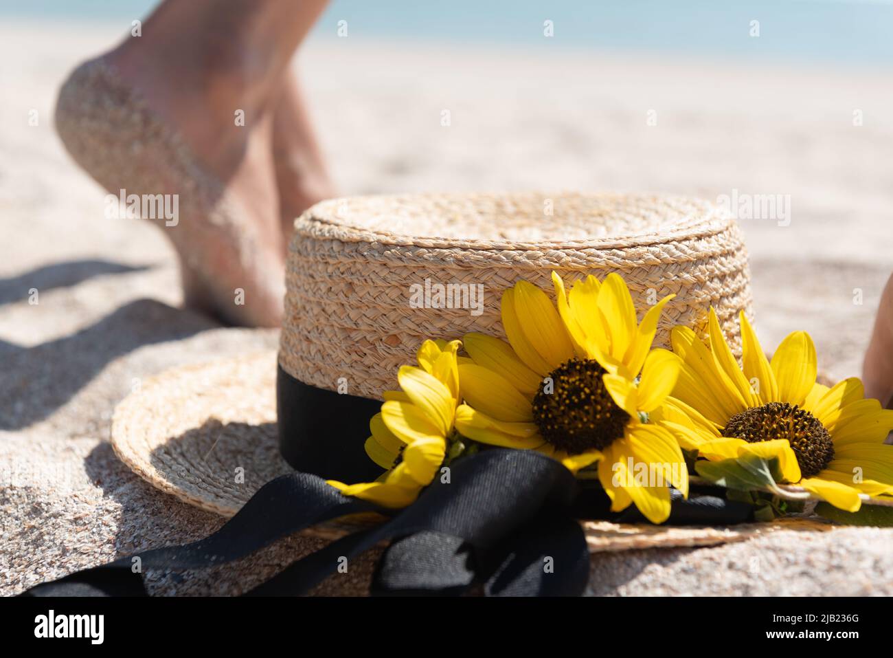 Blurred legs of a woman walking on the beach sand in the background with a straw hat and flowers close-up. Summer rest concept. Stock Photo