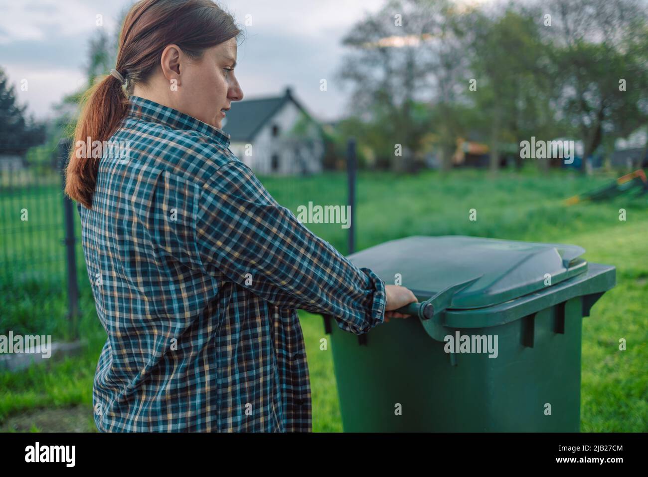 European 20s girl take out the trash can sorting garbage near a home. Female transports a green container with garbage to the backyard Stock Photo