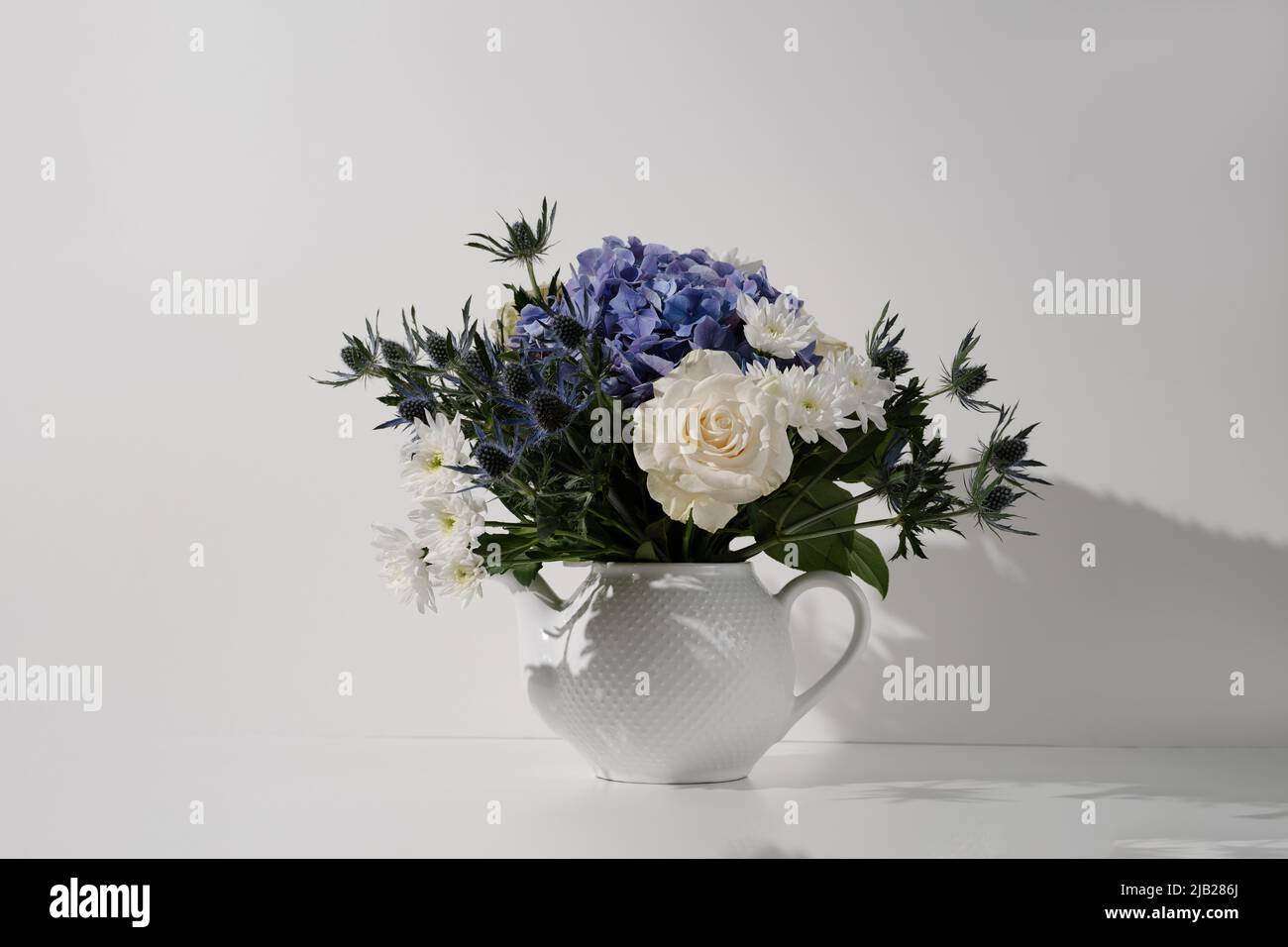 Flower bouquet with rose hydrangea chrysanthemum eryngium in a teapot on white background in sunlight. Modern hipster composition cottagecore sideview Stock Photo