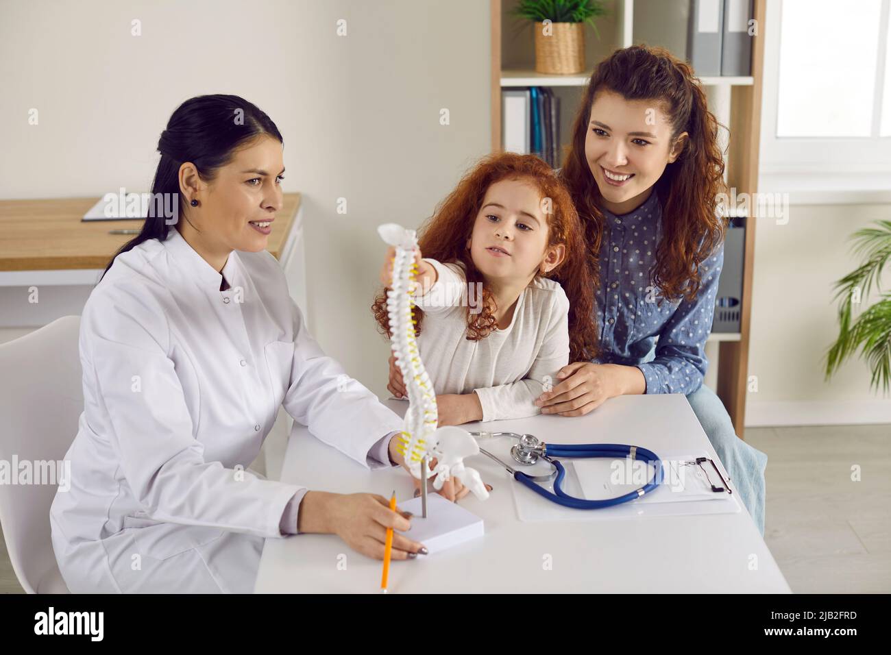Little cute girl examines anatomical model of spine who is standing on table in doctor's office. Stock Photo